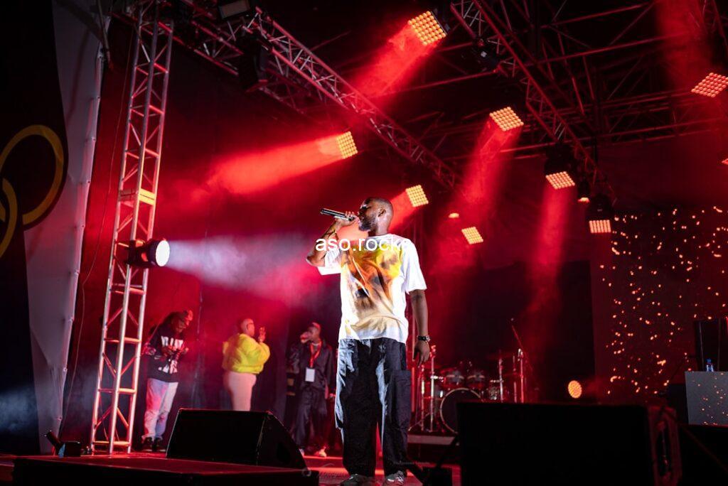 Music artist performing on stage under red lights at an outdoor concert with a large crowd
